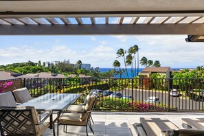 A covered outdoor area with a table and chairs, a view of a landscaped garden with palm trees, and a distant view of the ocean. - Wailea Elua, #2106 2 Bedroom Condo by RedAwning (Kihei)