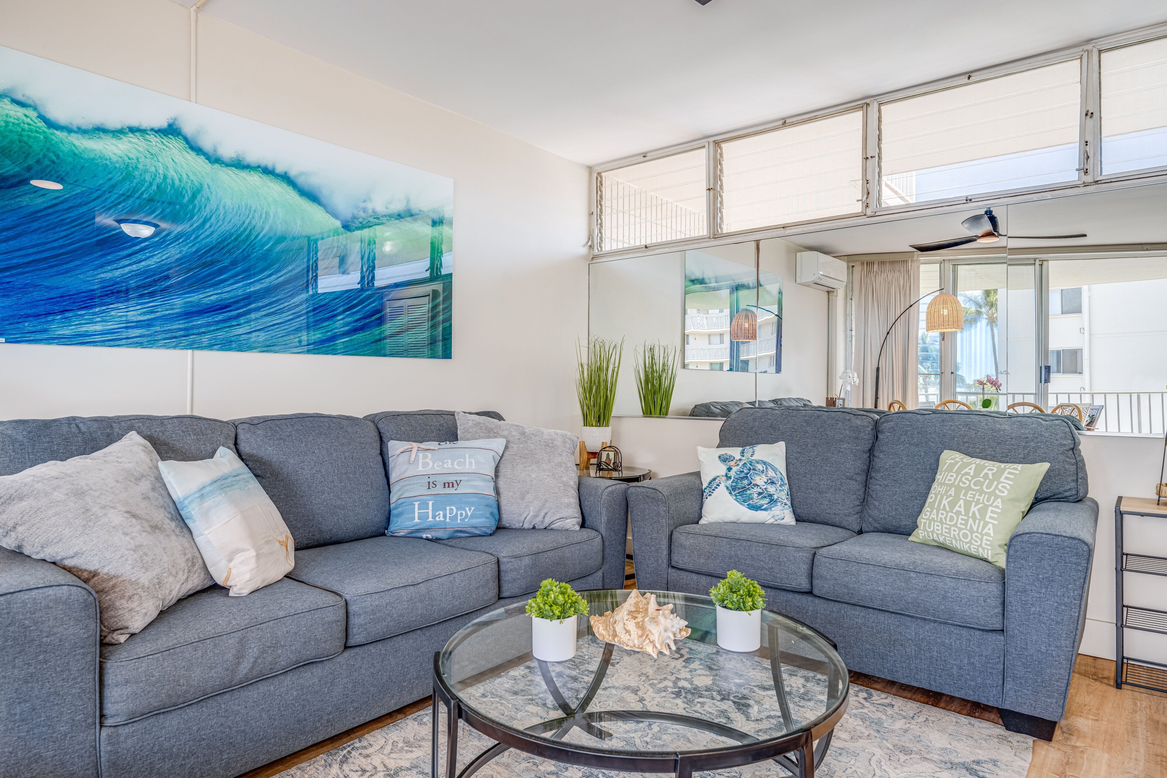 A modern living room with grey sofas, a glass coffee table, and a large wave-themed painting.
