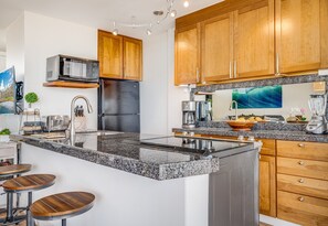 A modern kitchen with granite countertops, stainless steel appliances, wooden cabinets, and a central island with bar stools. - Kamaole Nalu, #502^ 2 Bedroom Condo by RedAwning (Kihei)