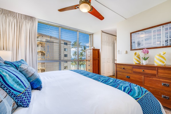 A bedroom with a large bed, a ceiling fan, a dresser, and a view of a building through a window.
