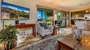 A modern living room with a large TV, comfortable seating, a kitchen area, and a view of the outdoors.