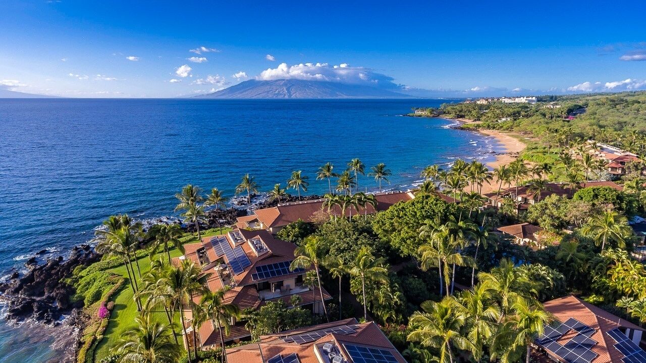 Um resort costeiro com vários edifícios, palmeiras e vista para o oceano e uma montanha ao longe.