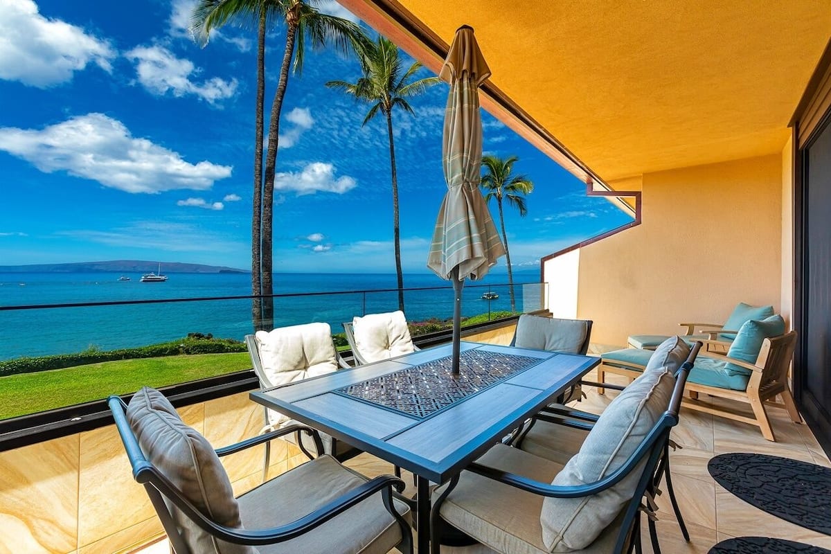 Ein Balkon mit Tisch und Stühlen, der Blick auf einen Strand mit Palmen und einem strahlend blauen Himmel bietet.
