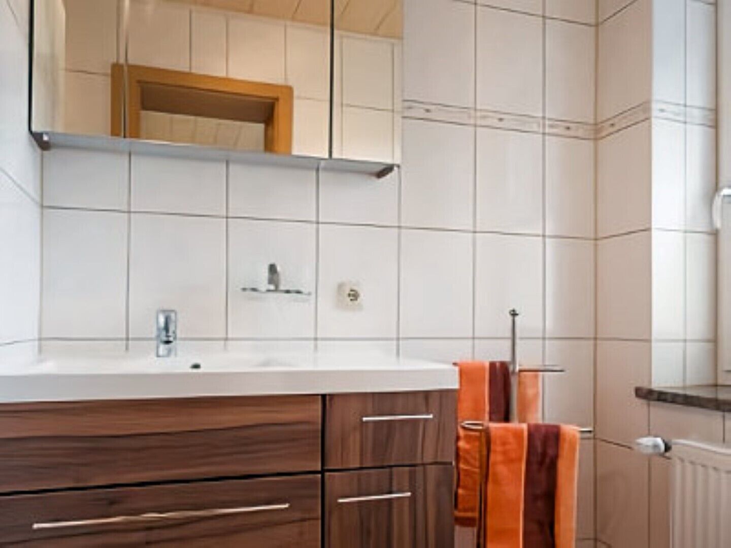 Un baño con pared de azulejos blancos, un tocador de madera con espejo, un lavabo y un perchero para toallas.