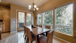 Un comedor con una gran mesa de madera y sillas de mimbre, ventanas amplias con vista a árboles y una araña.