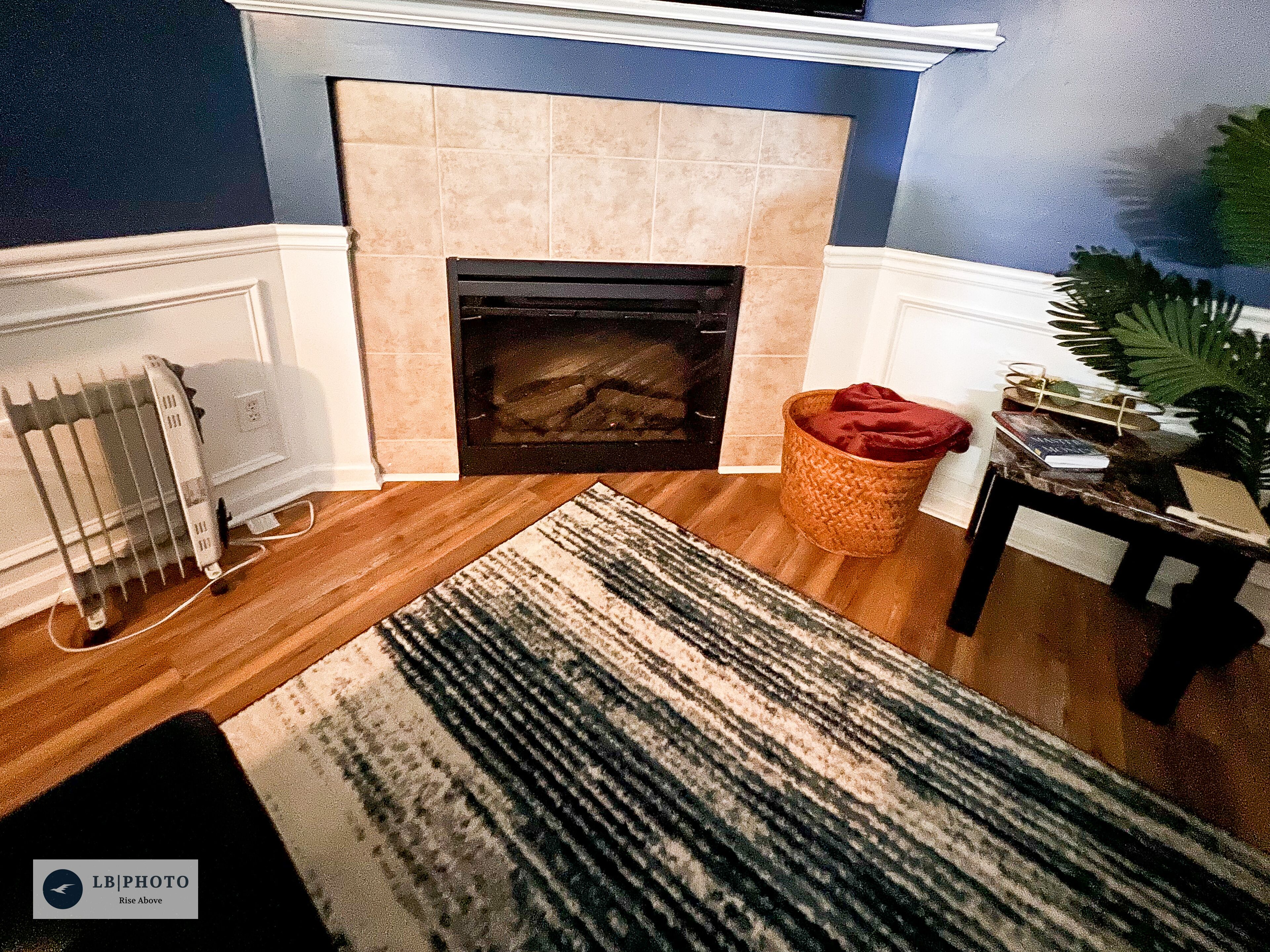 A living room with a fireplace, a radiator, a rug, a table with a plant, and a basket with red items.