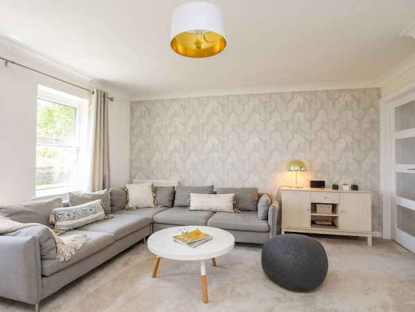 Un salon moderne avec un canapé d’angle gris, une table basse ronde et un pouf gris. On y trouve un meuble blanc sur lequel est posée une lampe, ainsi qu’un papier peint à motifs sur les murs.