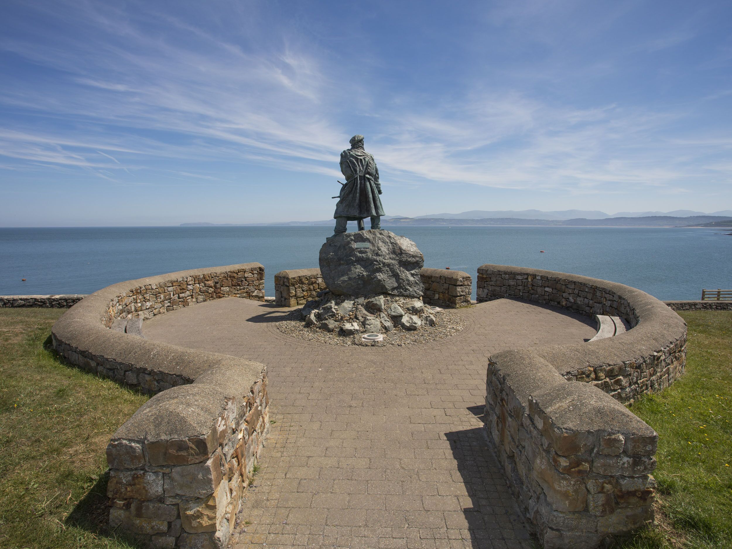 Una estatua sobre un pedestal de piedra con un muro circular de piedra y un camino pavimentado que conduce hacia ella.