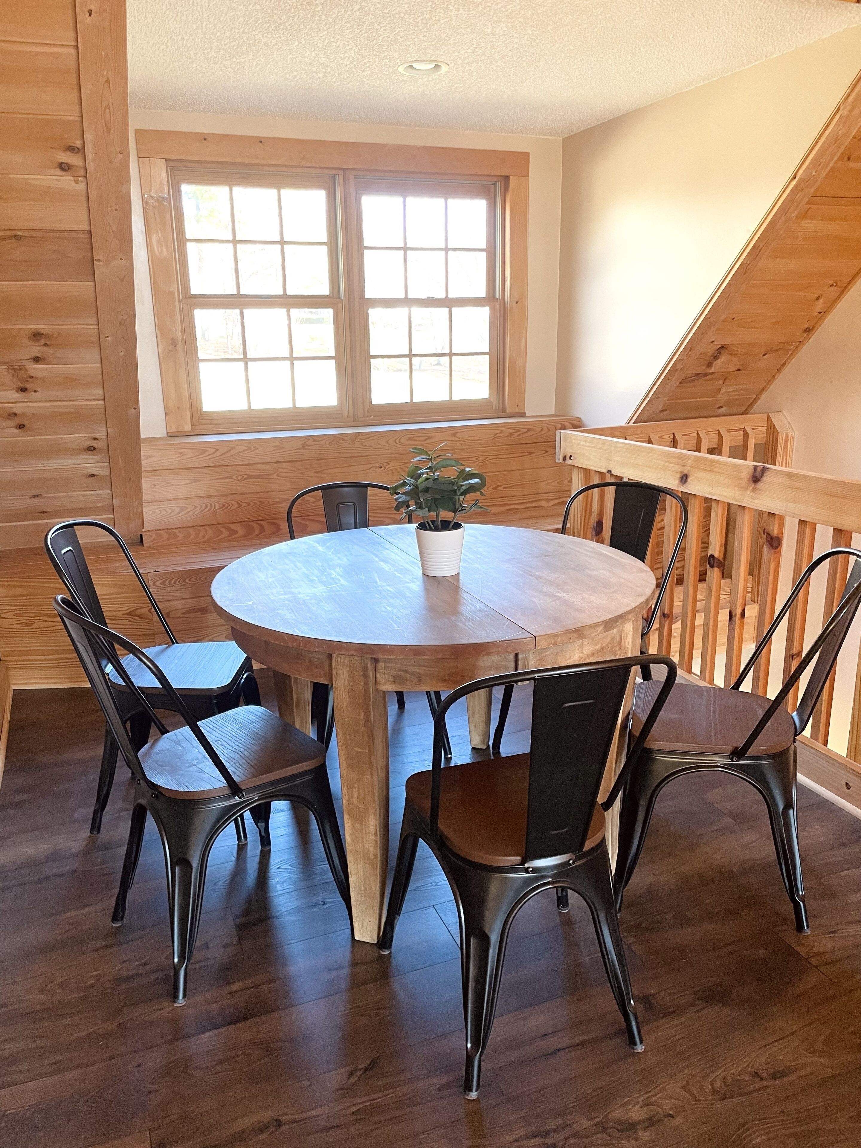 Une table de salle à manger ronde en bois, accompagnée de quatre chaises en métal, d’une plante en pot et d’une fenêtre donnant sur l’extérieur.