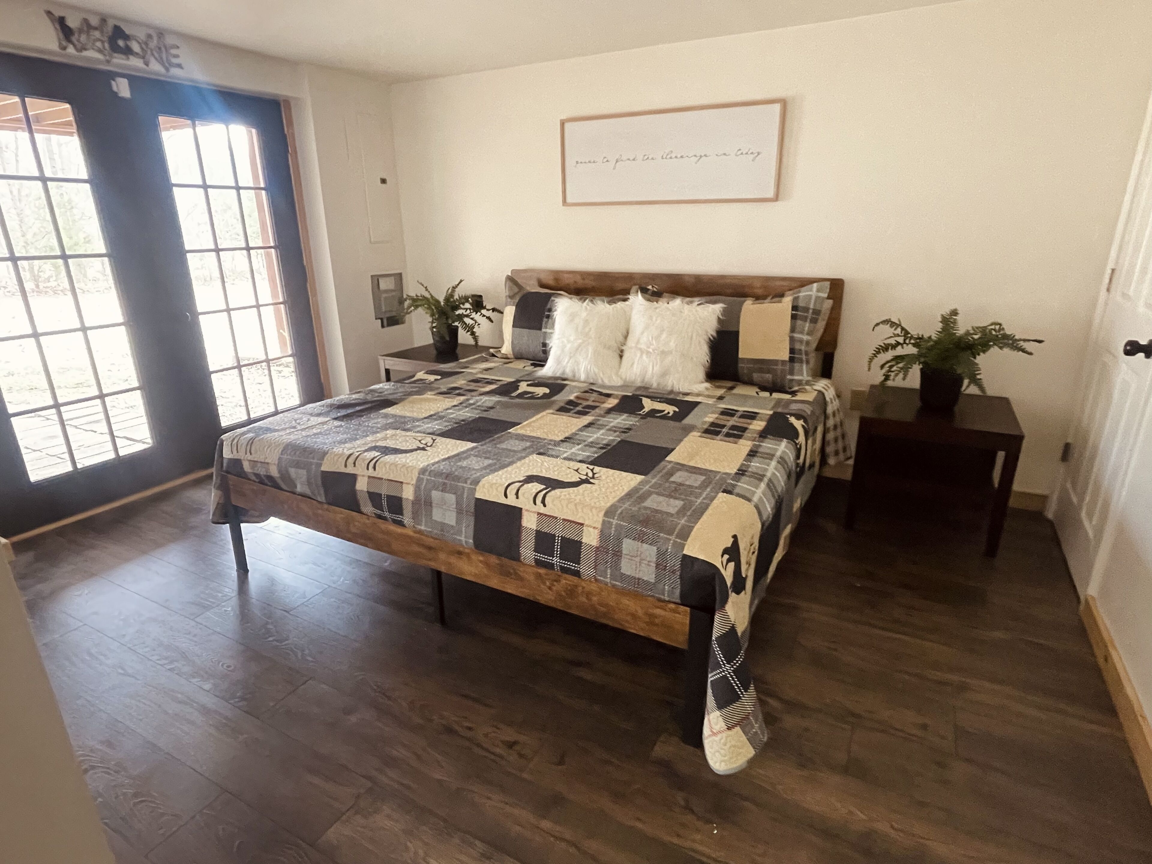 A bedroom with a large bed, a wooden headboard, a quilt, two bedside tables with lamps, a potted plant, and a door with a window.