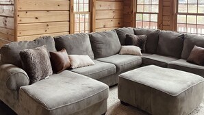 A cozy log cabin interior with a large sectional sofa, two windows, and a wooden ceiling.