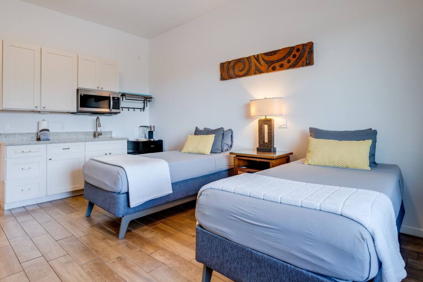 A compact hotel room with two single beds, a kitchenette, and a small bedside table with a lamp.