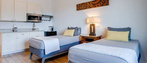 Une chambre d’hôtel compacte comprenant deux lits simples, une kitchenette et une petite table de chevet avec une lampe.