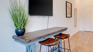 A modern room with a wooden bench, two wooden stools, a potted plant, a flat-screen TV mounted on the wall, and a framed picture. - Unit 22 Waena Inn - Maui Private Suite (Wailuku)