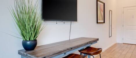 Una habitación moderna con un banco de madera, dos taburetes de madera, una planta en maceta, un televisor de pantalla plana montado en la pared y un cuadro enmarcado.