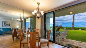 A dining area with wooden chairs and a glass table, a chandelier, and a view of a lush garden and ocean. - Kihei Beach, #107 1 Bedroom Condo by RedAwning (Kihei)