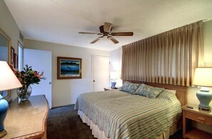 A hotel room with a bed, a ceiling fan, a window with curtains, a bedside table with a lamp, and a painting on the wall. - Kihei Beach, #405 1 Bedroom Condo by RedAwning (Kihei)