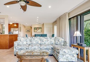 A living room with a sectional sofa, a glass coffee table, a wicker chair, a ceiling fan, recessed lighting, a kitchen area, and a large window with curtains. - Wailea Elua, #2501 2 Bedroom Condo by RedAwning (Kihei)