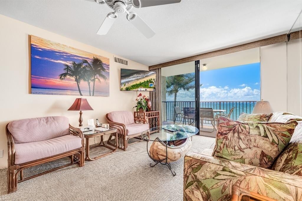 A room with a sofa, armchair, and a glass table. There is a ceiling fan, a painting of a sunset, and a view of the ocean through large windows.