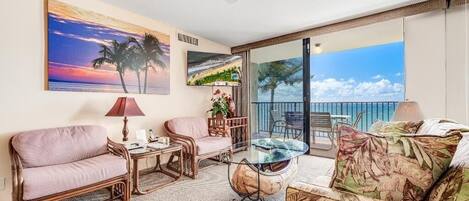 A room with a sofa, armchair, and a glass table. There is a ceiling fan, a painting of a sunset, and a view of the ocean through large windows.