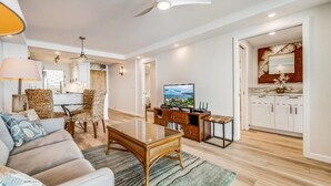 A modern living room with a sofa, dining table, and chairs, a kitchen with white cabinets, and a ceiling fan. - Royal Mauian, #517 2 Bedroom Condo by RedAwning (Kihei)