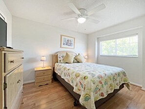Une chambre à coucher comprenant un lit, des tables de chevet, une commode, un ventilateur de plafond, une fenêtre et un tableau encadré au mur.
