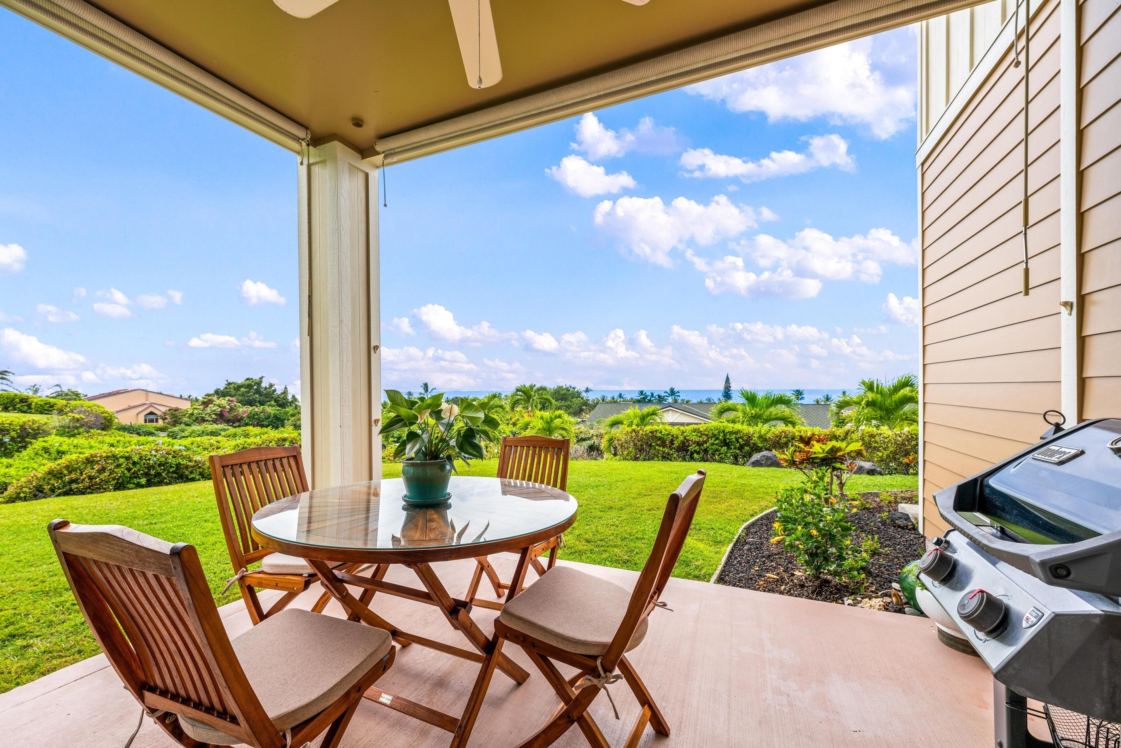 A covered patio with a round table and chairs, a grill, and a view of a well-maintained lawn and distant buildings.