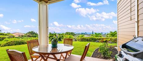 A covered patio with a round table and chairs, a grill, and a view of a well-maintained lawn and distant buildings.