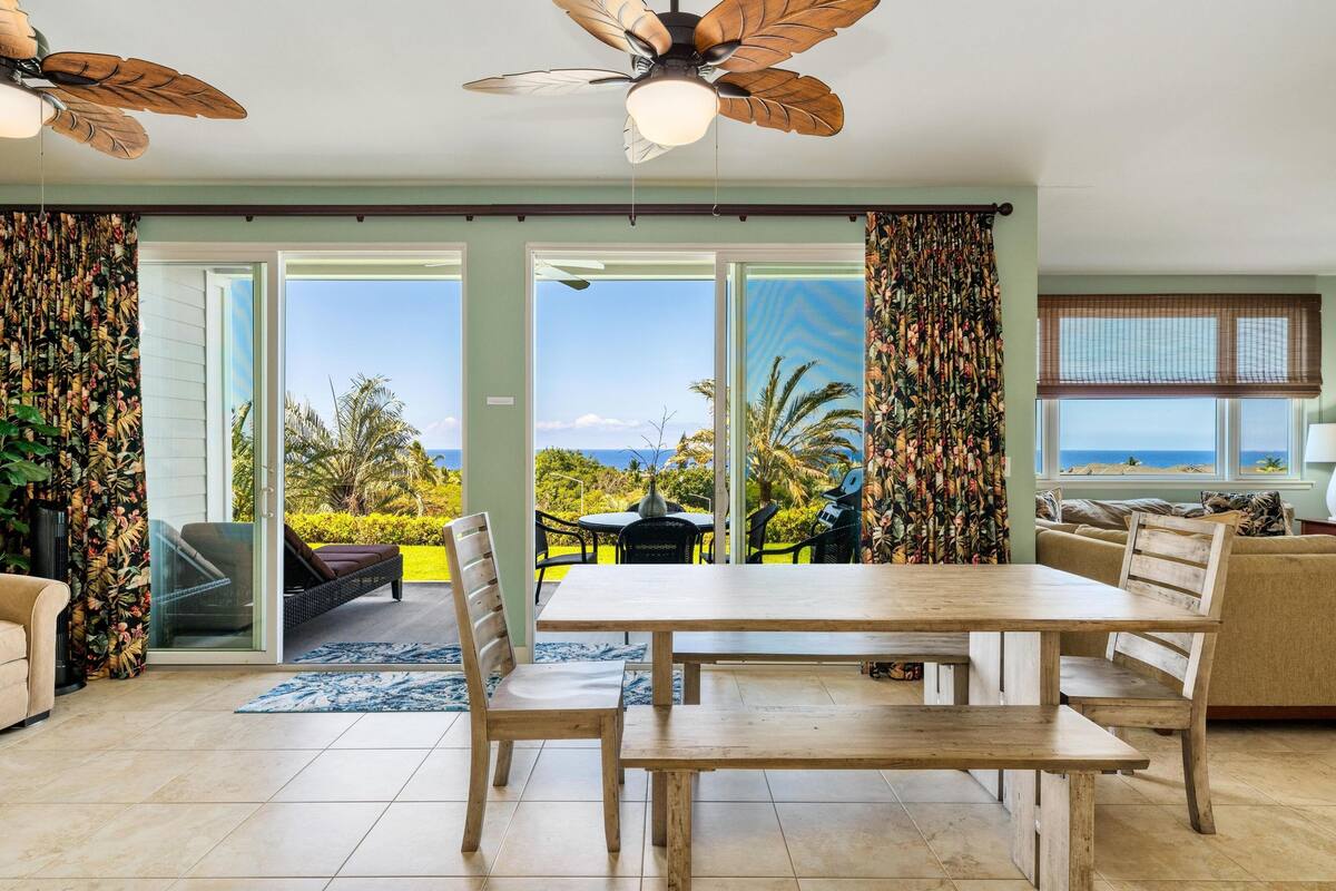 A spacious living room with a wooden dining table, two ceiling fans, and a large sliding glass door leading to a balcony with a view of the ocean.