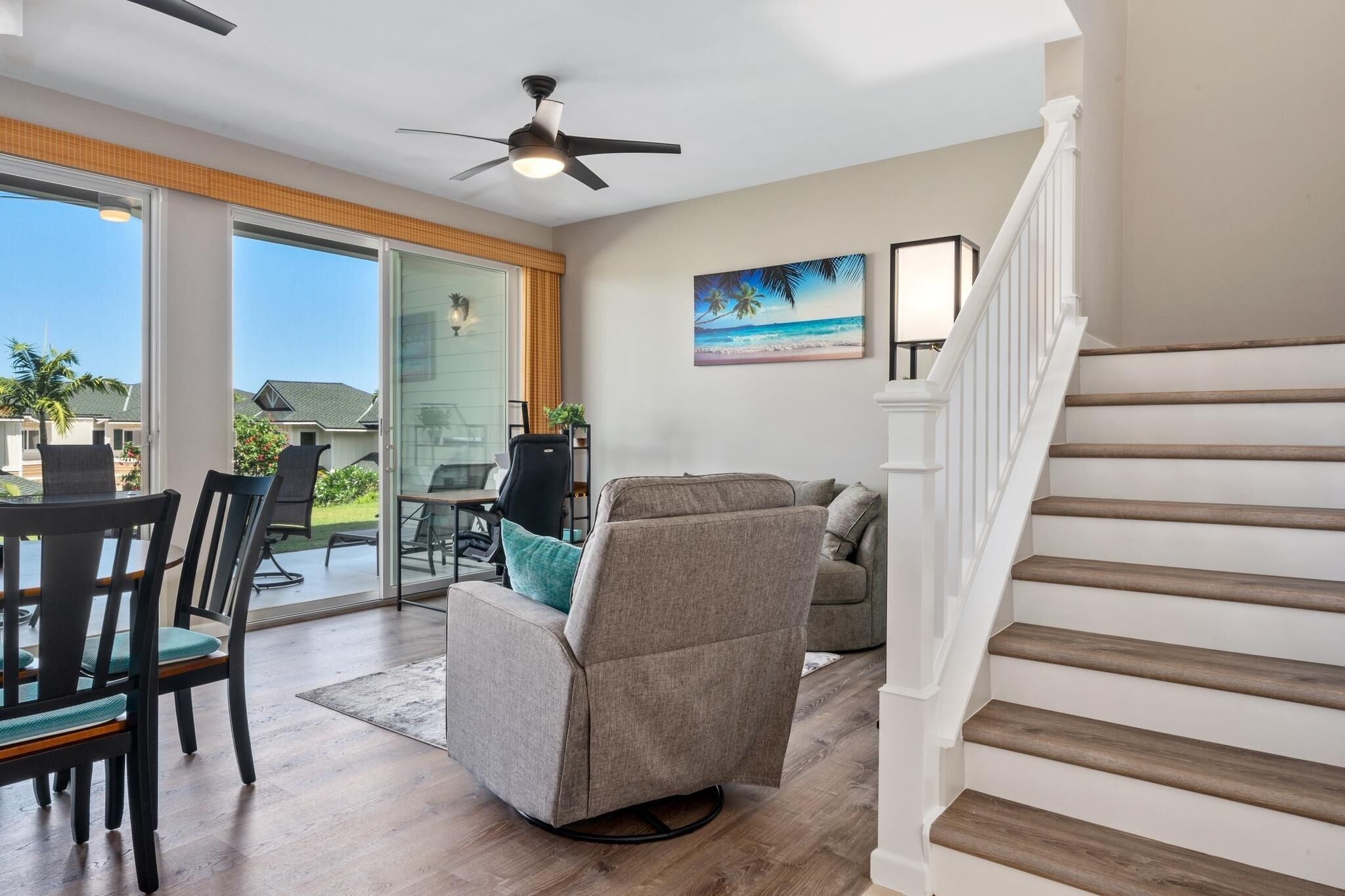 A modern living room with a sliding glass door leading to a patio, a ceiling fan, a painting of a beach scene, and a staircase.