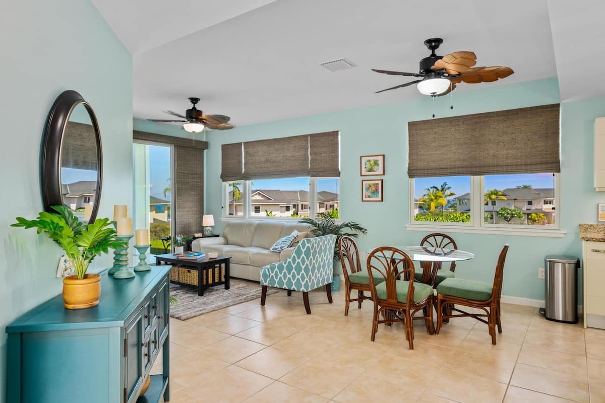 A living room with a sofa, dining table, chairs, a ceiling fan, and a view of neighboring houses through large windows.