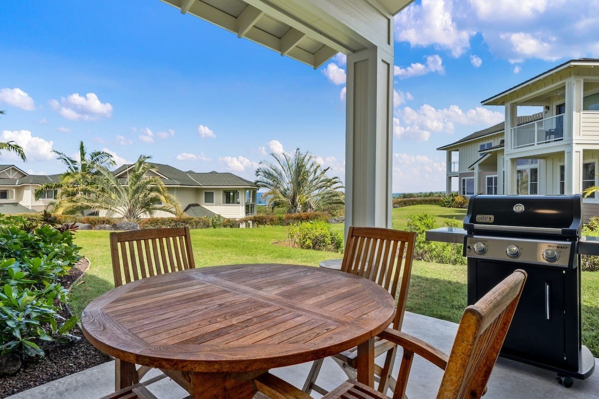 A patio with a round wooden table and chairs, a barbecue grill, and a view of a residential area with houses and greenery.