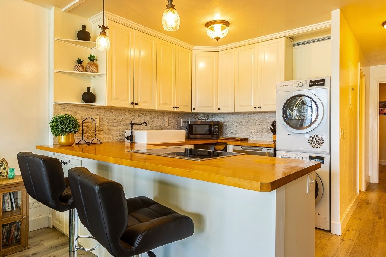 Une cuisine moderne avec des meubles blancs, un plan de travail en bois, un lave-linge encastré et un tabouret de bar.