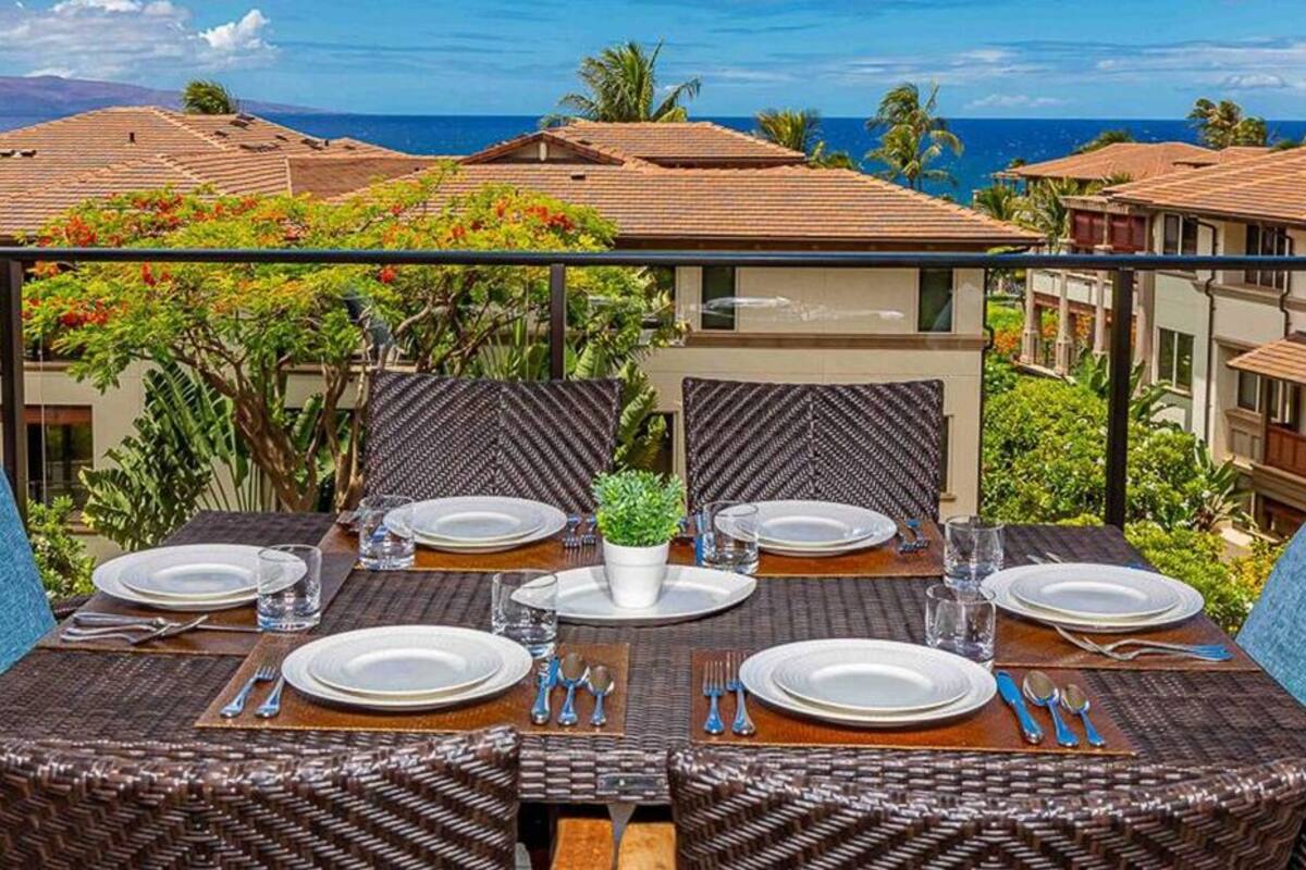 A dining area with wicker chairs and a table set for four, overlooking a coastal view with buildings and palm trees.