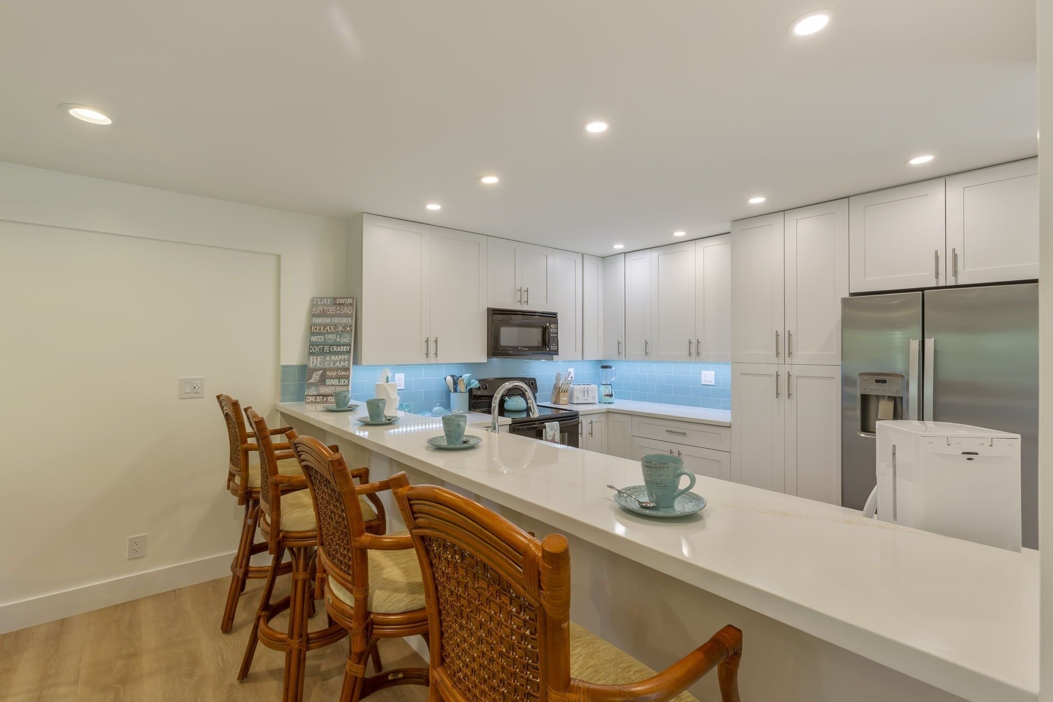 Una cocina moderna con armarios blancos, un refrigerador de acero inoxidable, un microondas y una encimera blanca alargada con taburetes de bar.