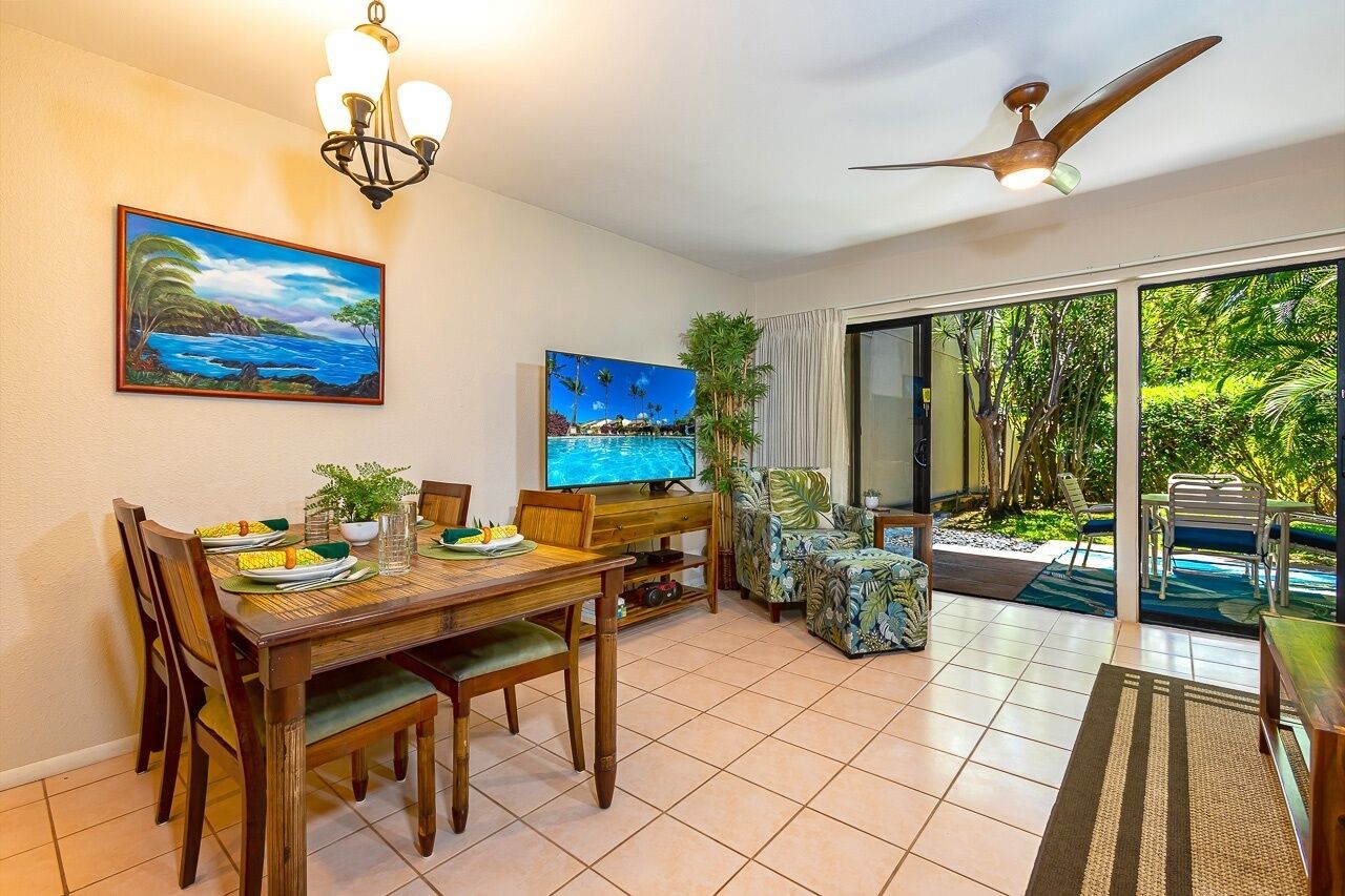 Uma sala de jantar com uma mesa de madeira, cadeiras e uma TV. Há um quadro na parede e vista para o verde lá fora.