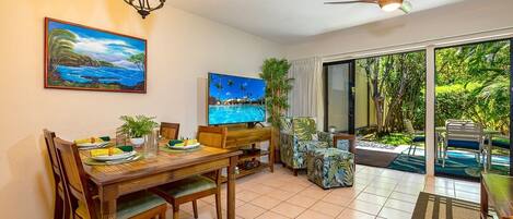 Uma sala de jantar com uma mesa de madeira, cadeiras e uma TV. Há um quadro na parede e vista para o verde lá fora.
