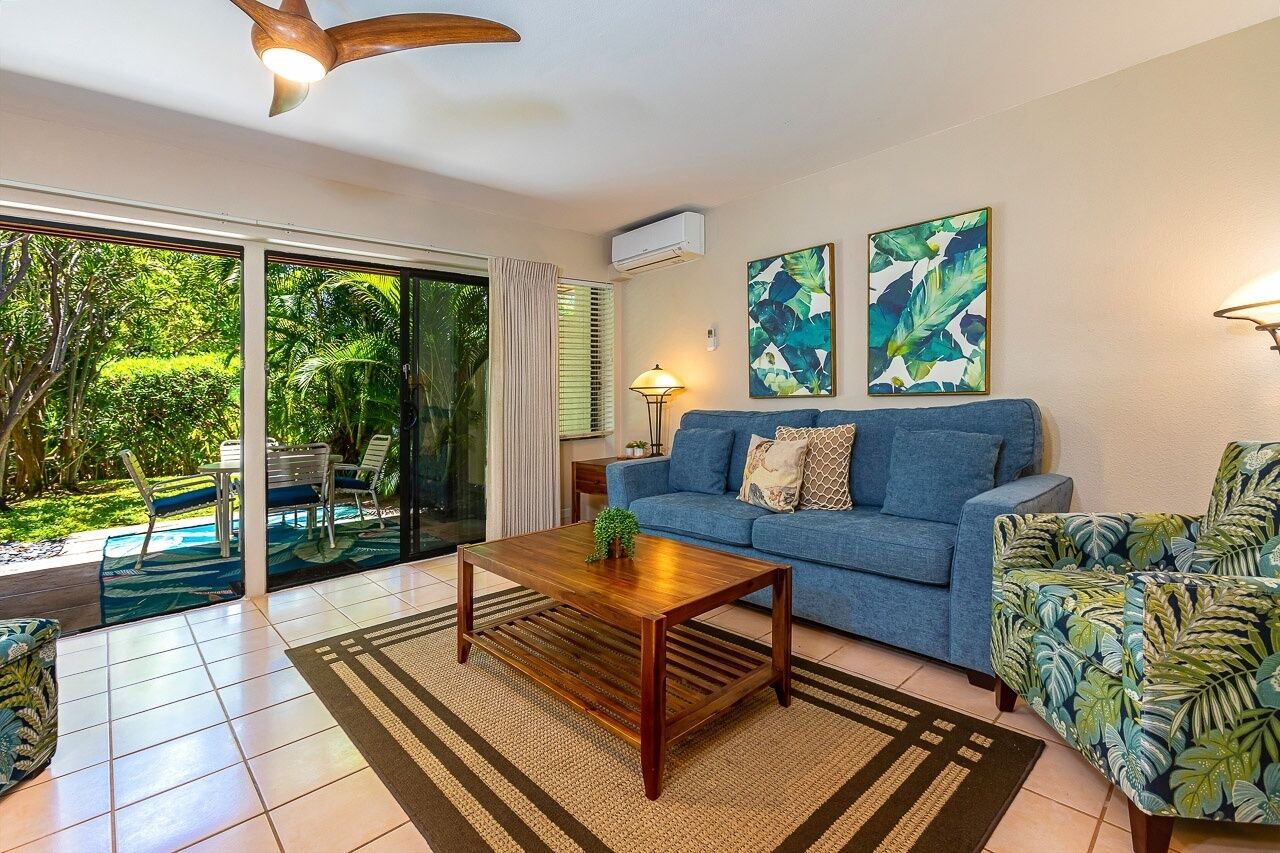 A living room with a blue sofa, a wooden coffee table, and a patterned armchair. There is a glass door leading to a garden.