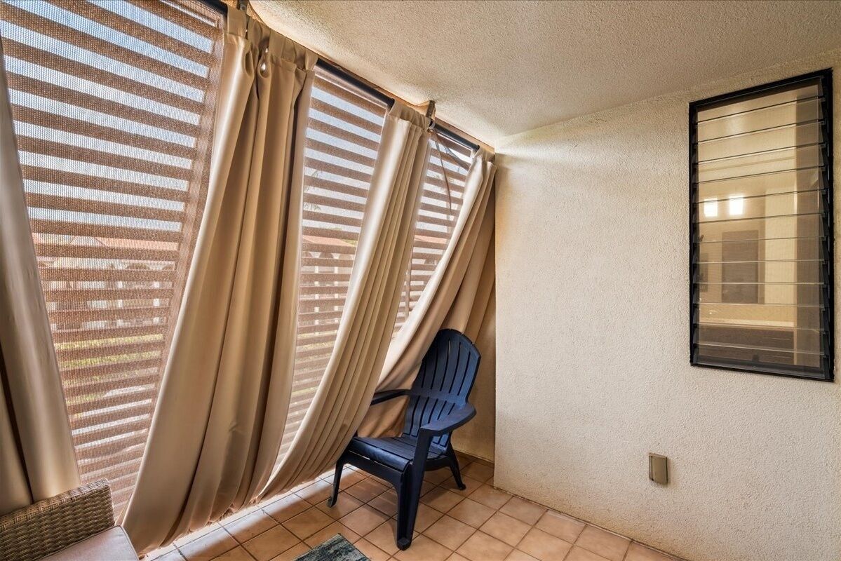 Habitación con una silla cerca de una ventana con cortinas color crema, piso de baldosas y una ventana con persianas horizontales fijadas a la pared.