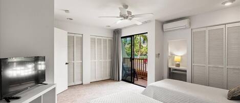 Habitación de hotel con cama, televisor, ventilador de techo y balcón con vistas a zonas verdes.