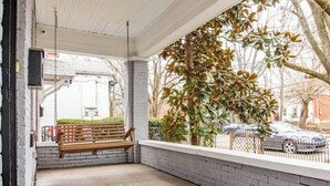 Un espacio de estar techado al aire libre con un banco, una cerca blanca y árboles de fondo.