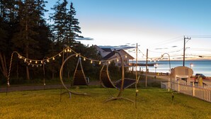 A grassy area with two swing chairs and string lights, a white picket fence, and a building with a covered structure in the background. - Navigateur Ste-Luce (Sainte-Luce)