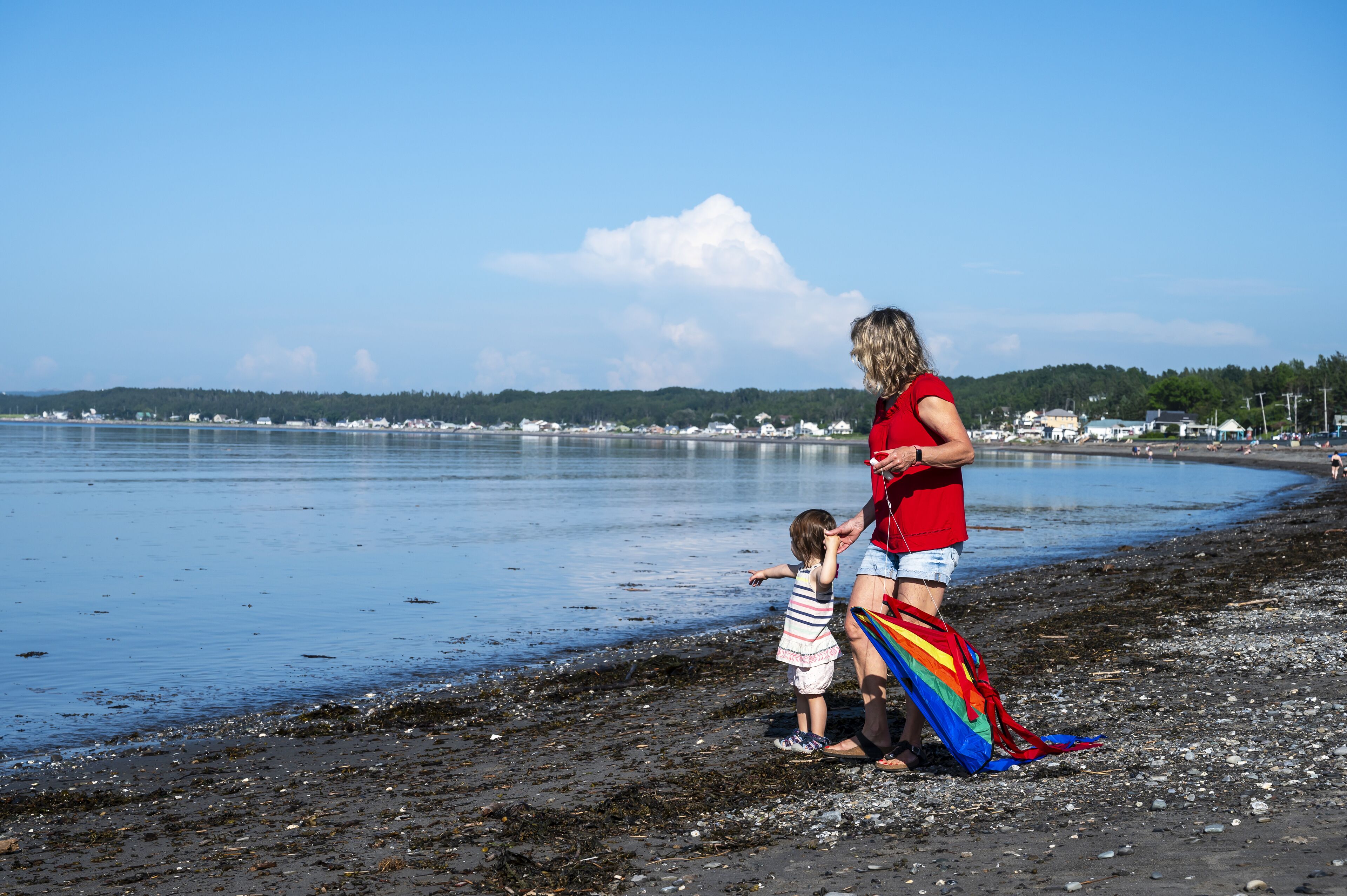 A beach scene with a woman and a child near the water, a rainbow kite, and a calm sea with buildings in the background.
