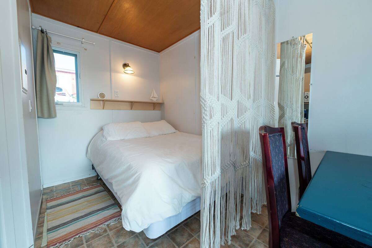 A bedroom with a bed, a window with curtains, a wall-mounted light, a macrame curtain, a wooden chair, and a blue table.