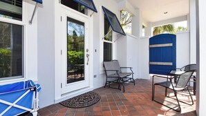 Ein überdachter Patio-Bereich mit einer weißen Tür, zwei Stühlen, einem Tisch und einem blauen Kinderwagen.