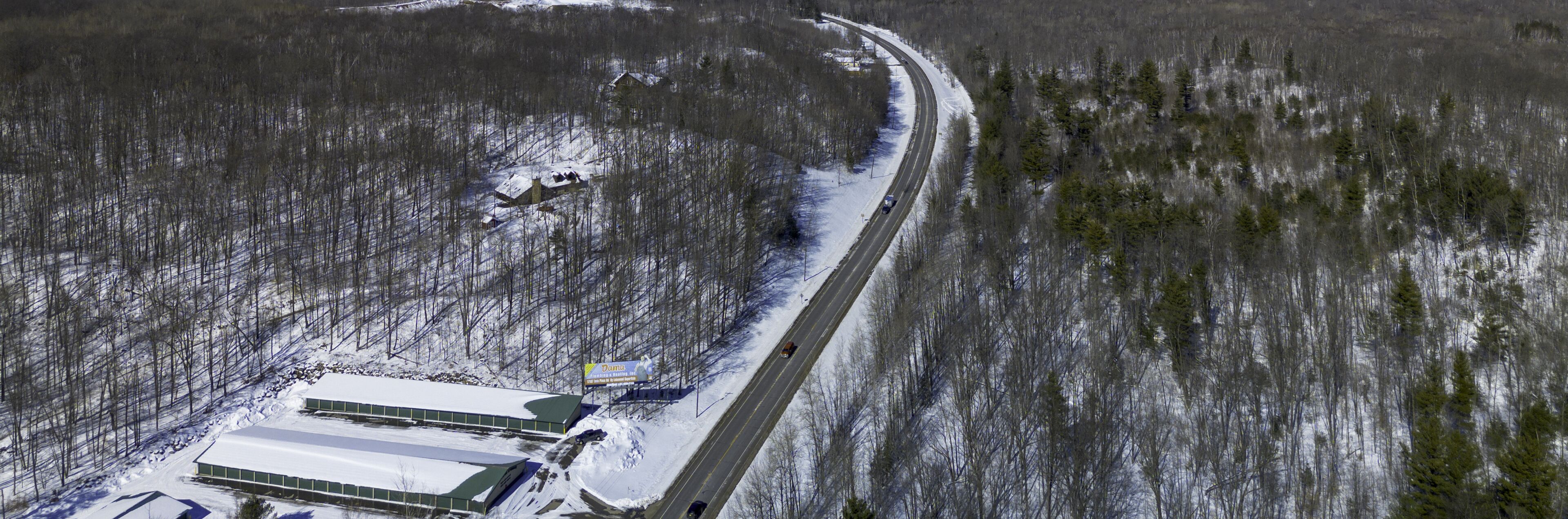 A snow-covered landscape with a road curving through a forest. There are buildings with snow on their roofs, and a billboard is visible.