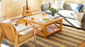A cozy living room with a wooden coffee table, a comfortable armchair, a sofa, a guitar, and a window with natural light.