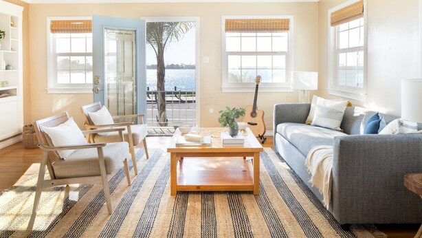A living room with a sofa, chairs, a coffee table, and a guitar. There are windows with blinds and a view of a body of water.