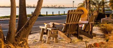 A beachside seating area with wooden chairs and a small table, overlooking a body of water with a distant city skyline.