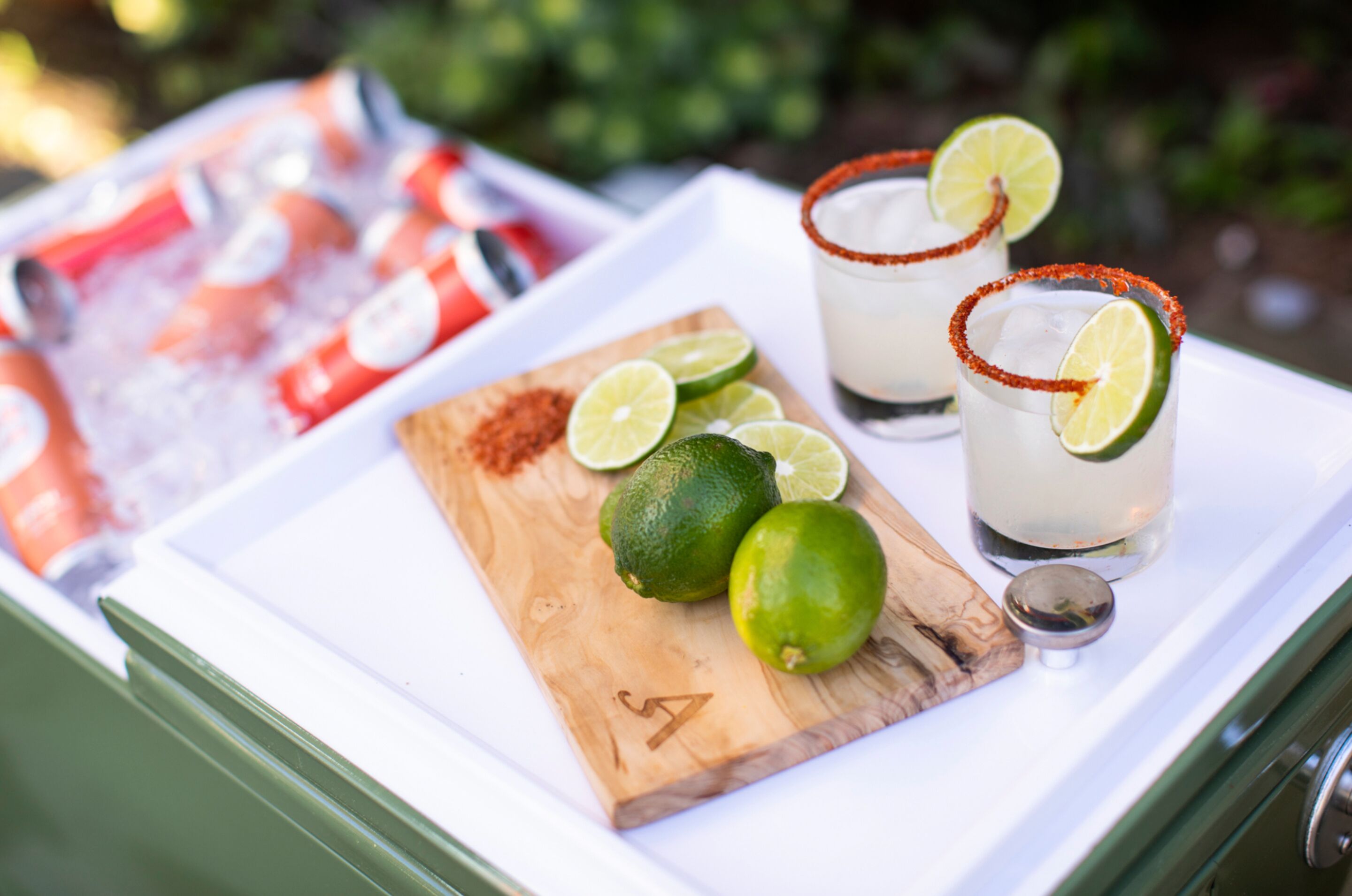 Un plateau avec deux margaritas décorées de quartiers de citron vert et de sel, une planche en bois avec des citrons verts et une glacière remplie de bouteilles.